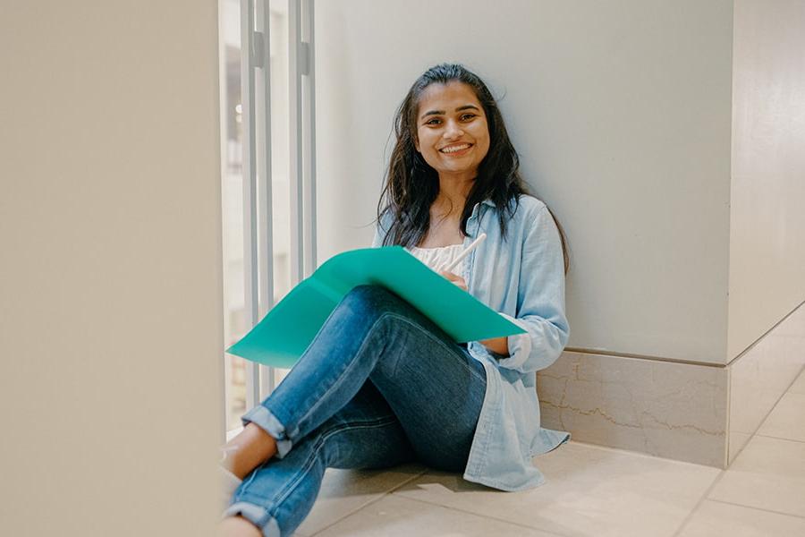 Nursing student sits on ground in campus center with laptop smiling.